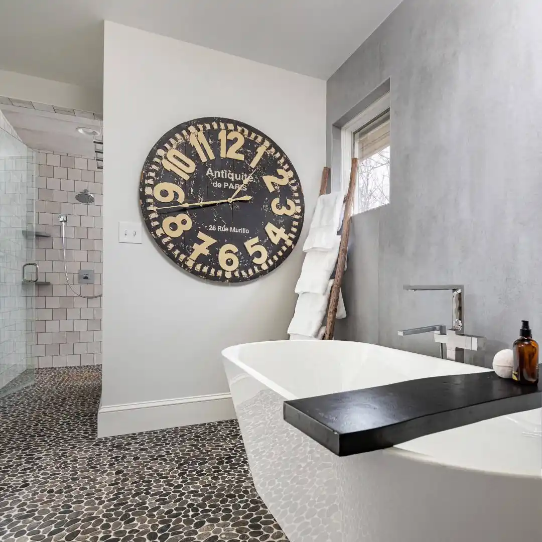 Modern bathroom with a freestanding white tub and large black and white wall clock, reminiscent of the elegance found in top landscaping services Delaware County PA offers. Pebble-style tiles cover the floor, complemented by a towel ladder rack. The shower area boasts white tiled walls and a sleek glass door.