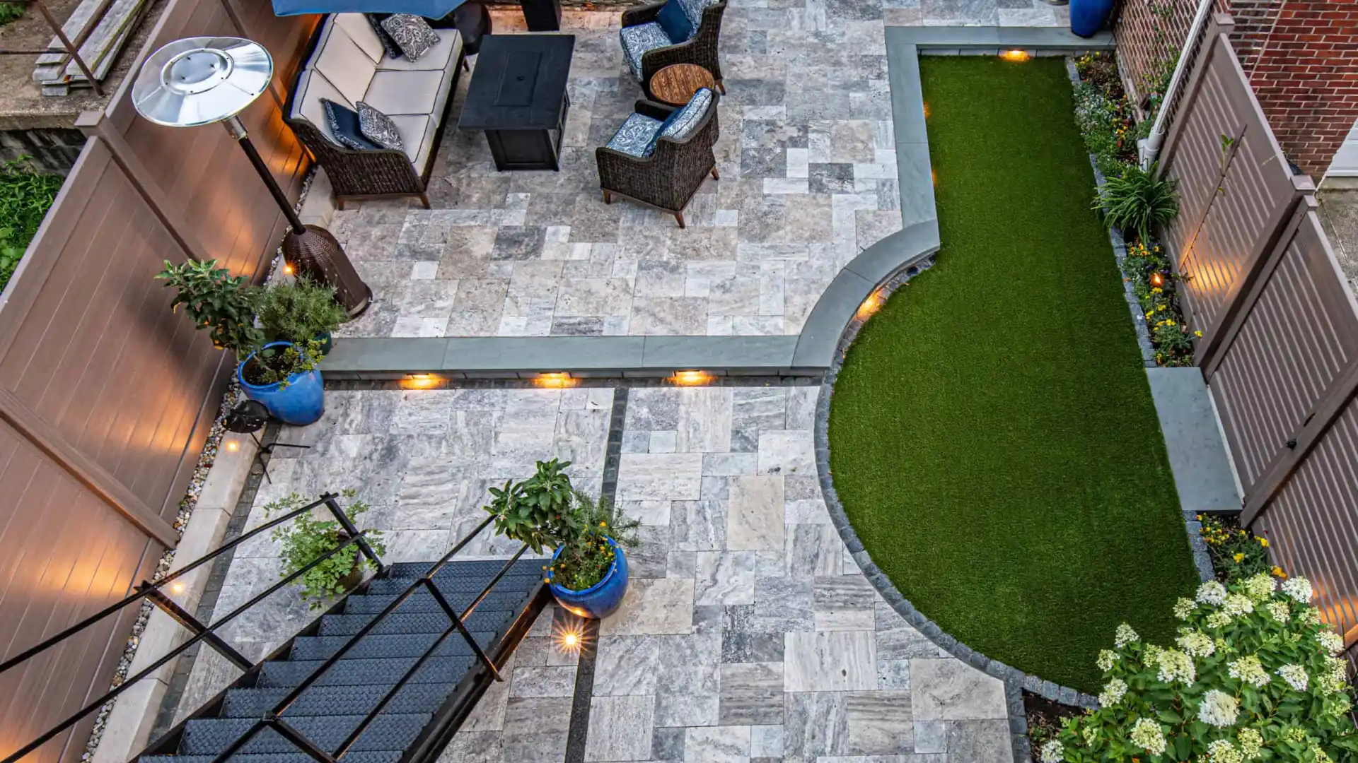 Aerial view of a modern patio in Delaware County, PA, with stone tiles, artificial grass, and cozy seating. Highlighted by a black metal staircase, blue planters, decorative lighting, and a dining set under a dark umbrella. Brick walls wrap around this charming outdoor living space.