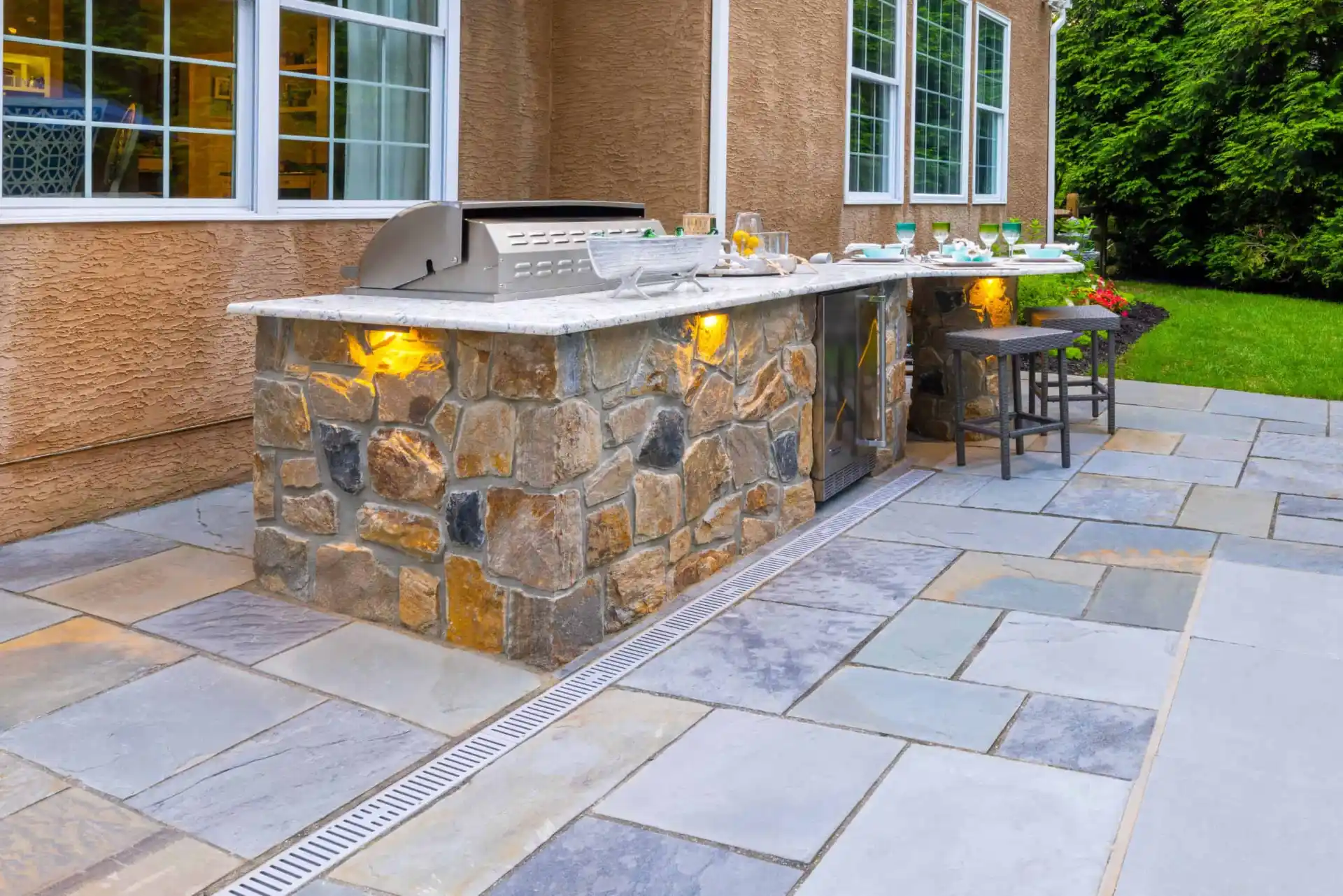 Outdoor kitchen with a stone countertop and grill set into a brick base. Two barstools are at the corner, nestled on a patio with multicolored stone tiles. Surrounded by lush greenery and a building with large windows, it's an ideal spot to enjoy outdoor living in Delaware County, PA.