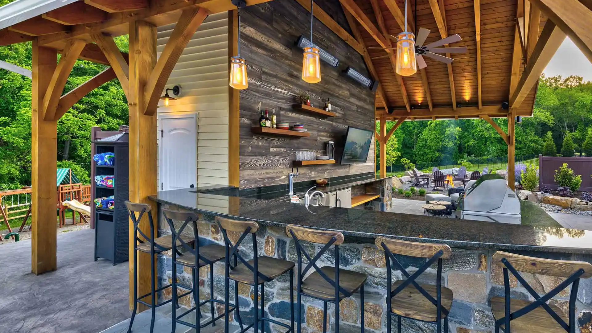 An outdoor bar with a wooden roof and stone counter, surrounded by metal high chairs, embodies the essence of outdoor living in Delaware County PA. Shelves stocked with bottles, a TV, and pendant lights add charm to the space, set against a lush backdrop of trees and an inviting seating area.
