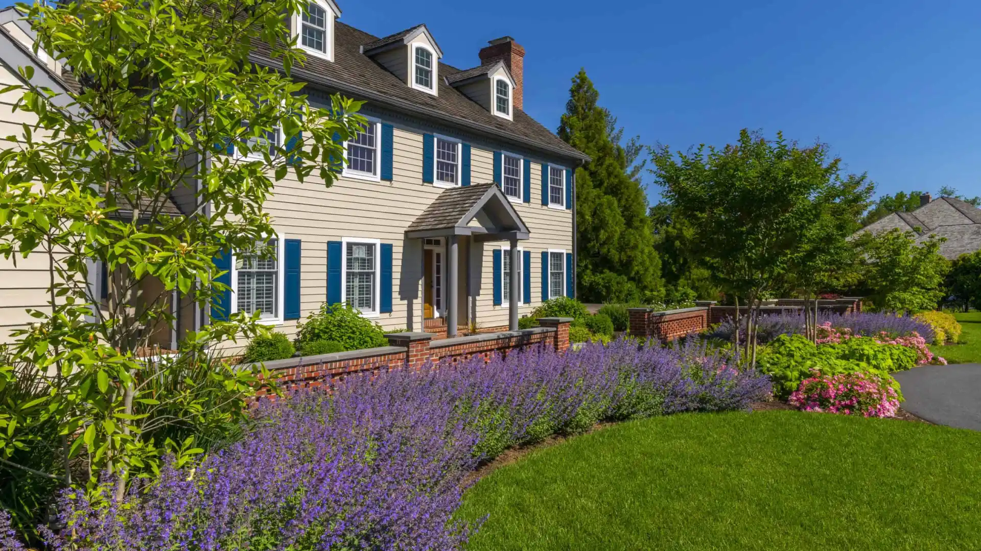 This charming two-story house, with beige siding and blue shutters, is nestled in lush greenery. Vibrant flowers and expertly designed hardscaping services in Delaware County, PA enhance its traditional appeal, complete with brick pathways under a clear blue sky.