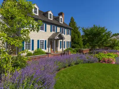 This charming two-story house, with beige siding and blue shutters, is nestled in lush greenery. Vibrant flowers and expertly designed hardscaping services in Delaware County, PA enhance its traditional appeal, complete with brick pathways under a clear blue sky.