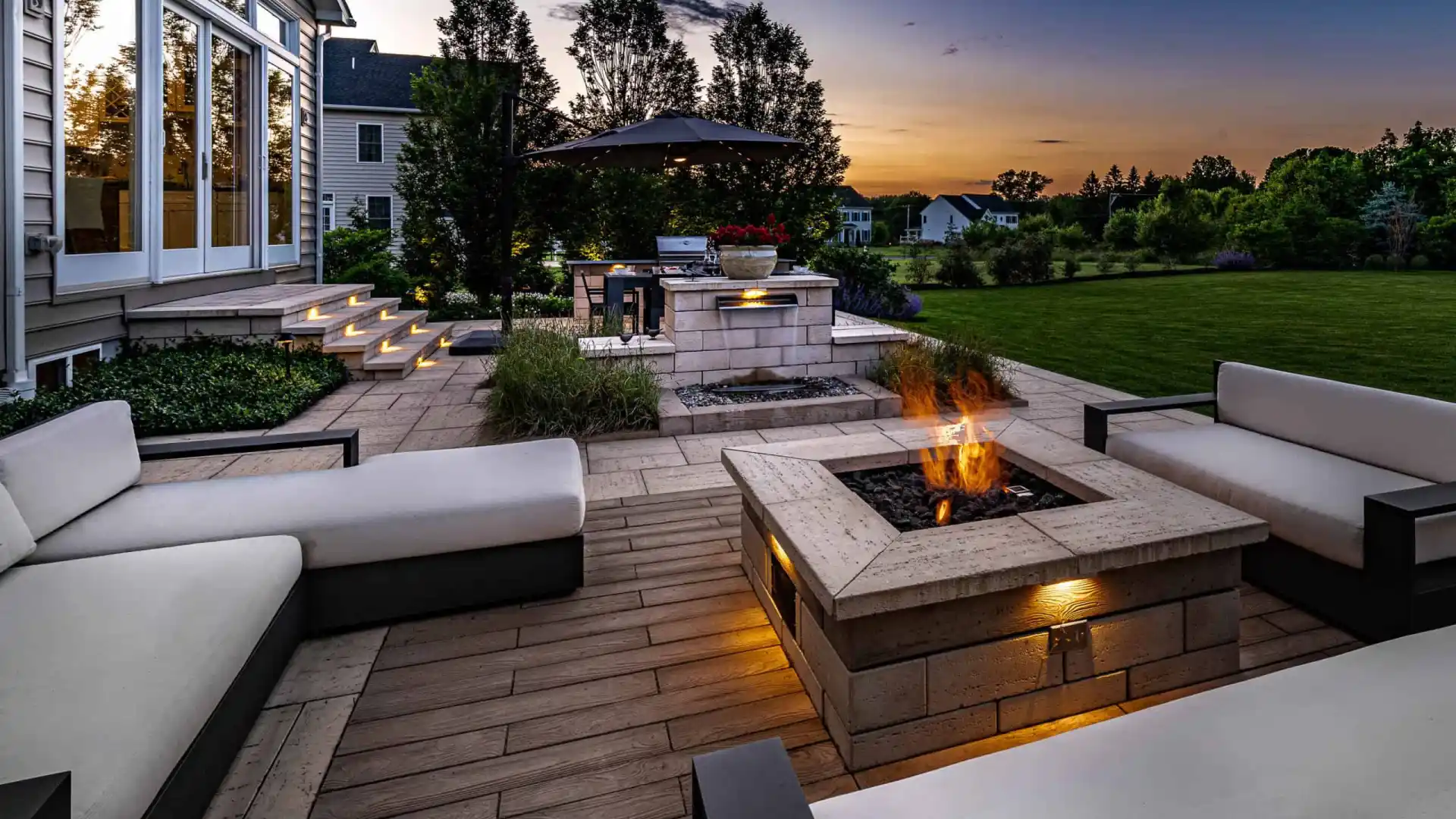 Outdoor living at its finest in Delaware County, PA, with cushioned seating around a lit fire pit. Steps with lighting guide you to the house, and a barbecue station with an umbrella is nearby. Overlooking a large lawn and garden beneath a sunset's warm glow, it's the perfect retreat.