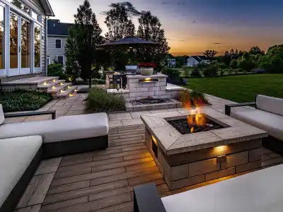 Outdoor living at its finest in Delaware County, PA, with cushioned seating around a lit fire pit. Steps with lighting guide you to the house, and a barbecue station with an umbrella is nearby. Overlooking a large lawn and garden beneath a sunset's warm glow, it's the perfect retreat.