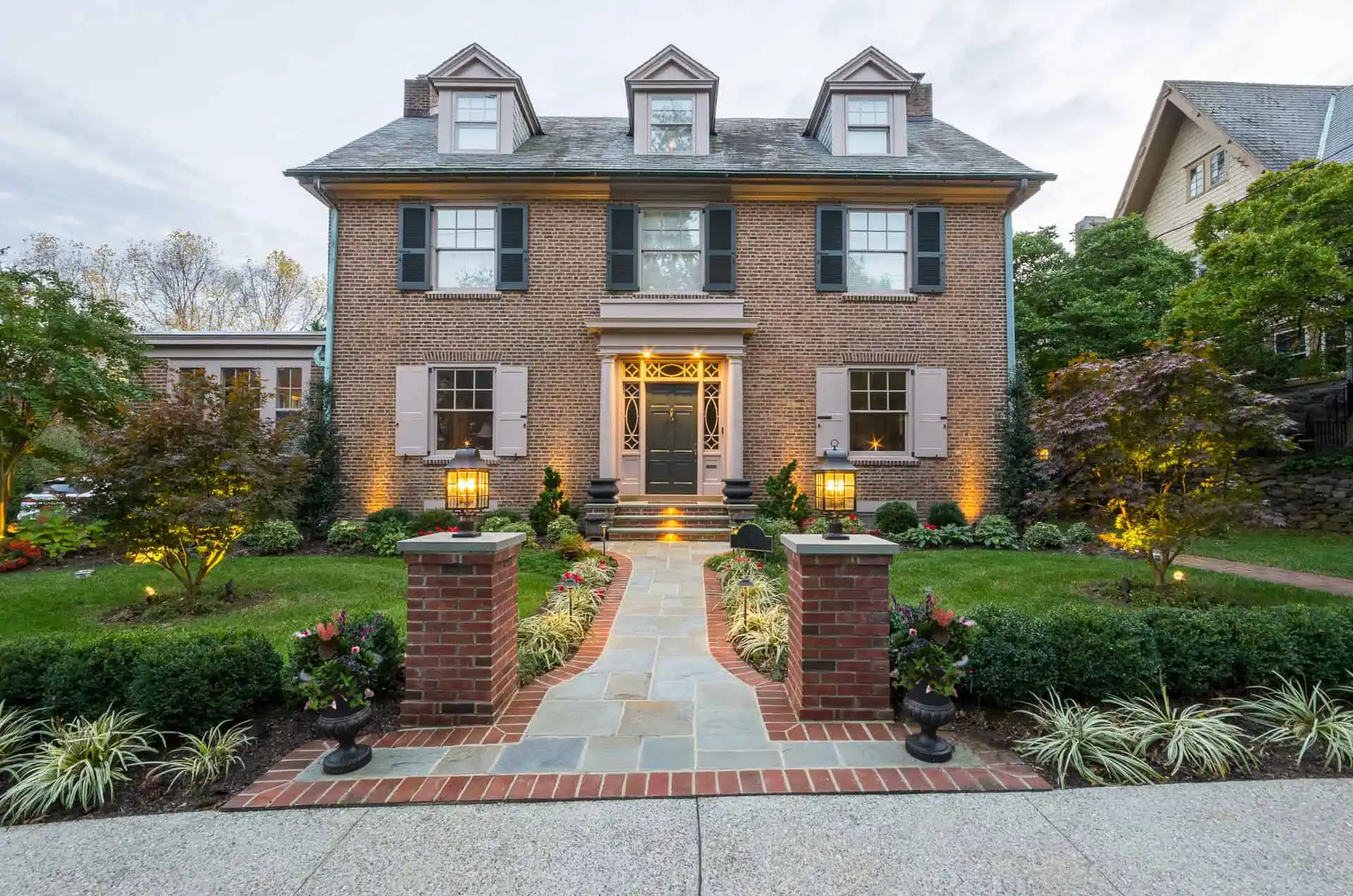 A stately two-story brick house with three dormer windows and a symmetrical layout features a landscaped front yard enhanced by top-tier landscaping services in Delaware County, PA. A stone path leads to the entrance, flanked by brick pillars and lanterns, with shrubs and trees arranged neatly around the yard.
