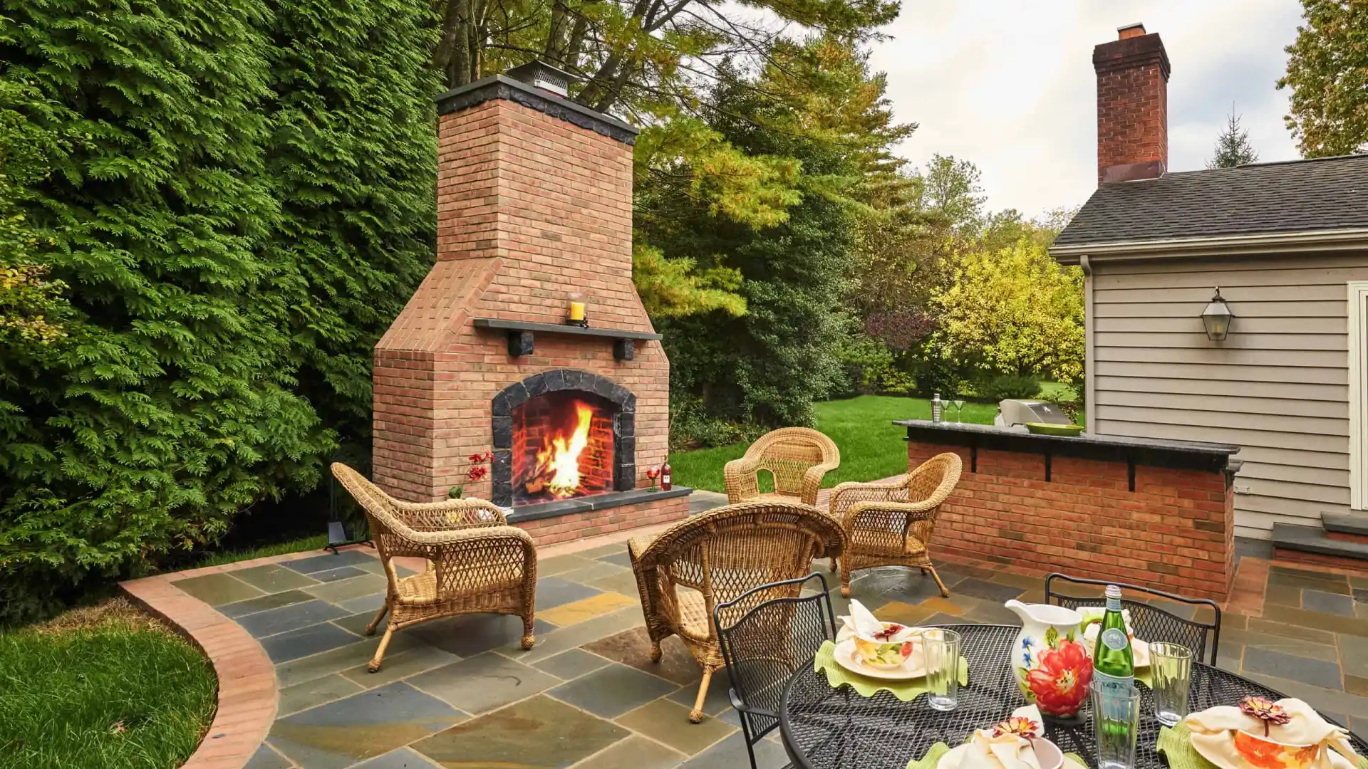 A patio featuring an inviting lit outdoor fireplace is surrounded by wicker furniture. A round metal table is elegantly set for four with plates, wine glasses, and a bottle. Lush greenery and trees from top-notch landscaping services in Delaware County PA create a serene backdrop.