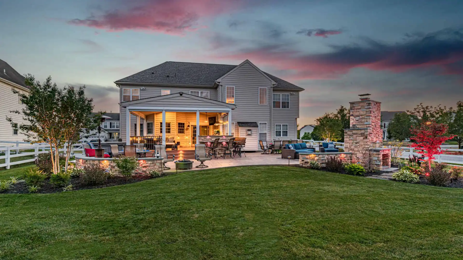 Backyard view of a large house at sunset in Delaware County, PA. The patio is illuminated, featuring outdoor seating, a fire pit, and a stone fireplace. There's expertly maintained landscaping with colorful plants and a dramatic sky with pink and blue hues overhead.