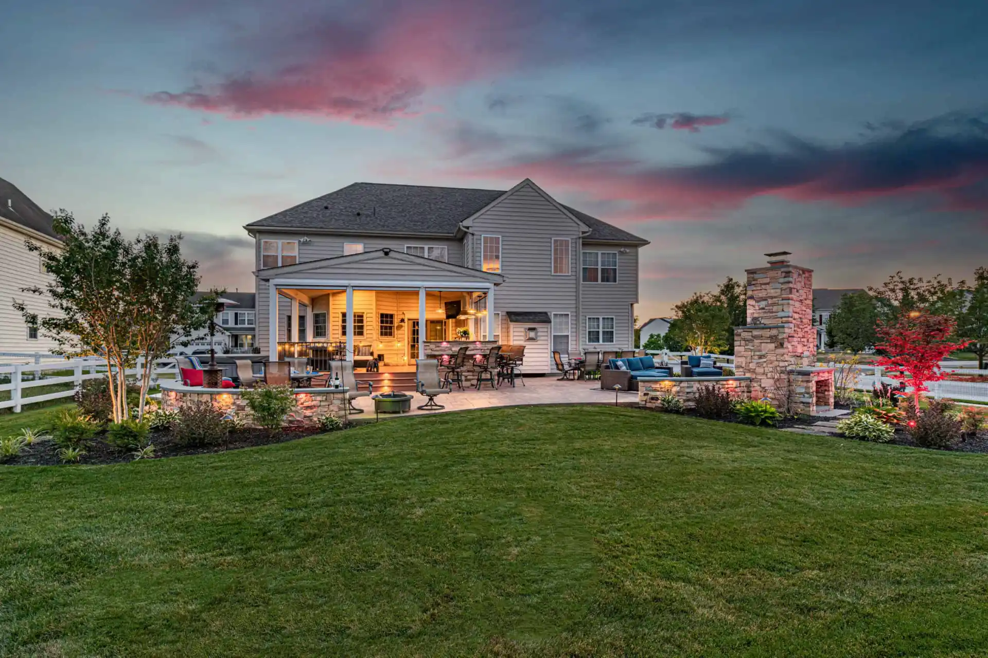 Backyard view of a large house at sunset in Delaware County, PA. The patio is illuminated, featuring outdoor seating, a fire pit, and a stone fireplace. There's expertly maintained landscaping with colorful plants and a dramatic sky with pink and blue hues overhead.