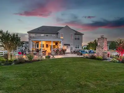 Backyard view of a large house at sunset in Delaware County, PA. The patio is illuminated, featuring outdoor seating, a fire pit, and a stone fireplace. There's expertly maintained landscaping with colorful plants and a dramatic sky with pink and blue hues overhead.