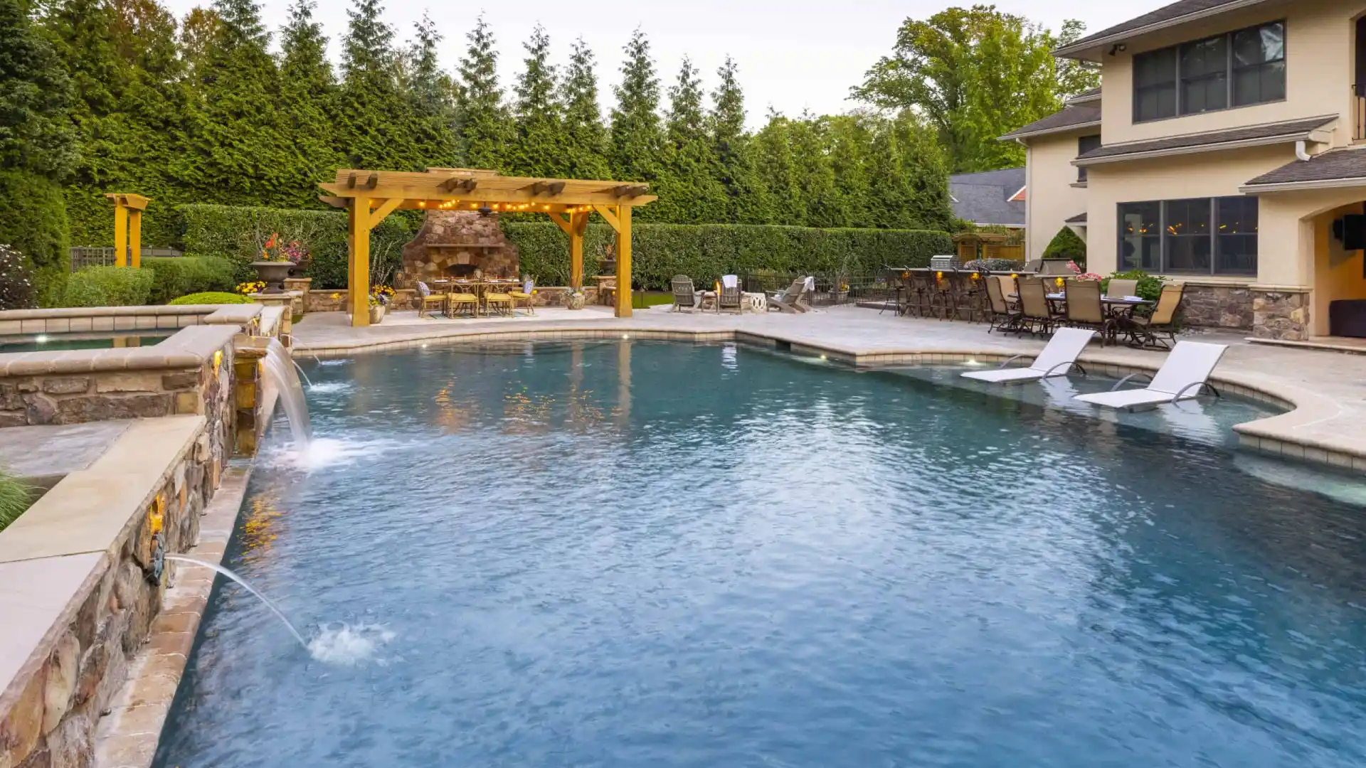 A luxurious backyard in Delaware County, PA, showcases a large swimming pool with water jets and stone decking. A wooden pergola with a fireplace and seating area accentuates the space, while lounge chairs partially submerged in the pool invite relaxation against a backdrop of expertly crafted hardscaping.