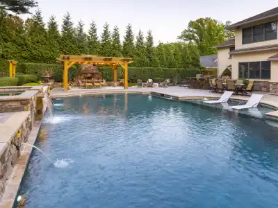 A luxurious backyard in Delaware County, PA, showcases a large swimming pool with water jets and stone decking. A wooden pergola with a fireplace and seating area accentuates the space, while lounge chairs partially submerged in the pool invite relaxation against a backdrop of expertly crafted hardscaping.