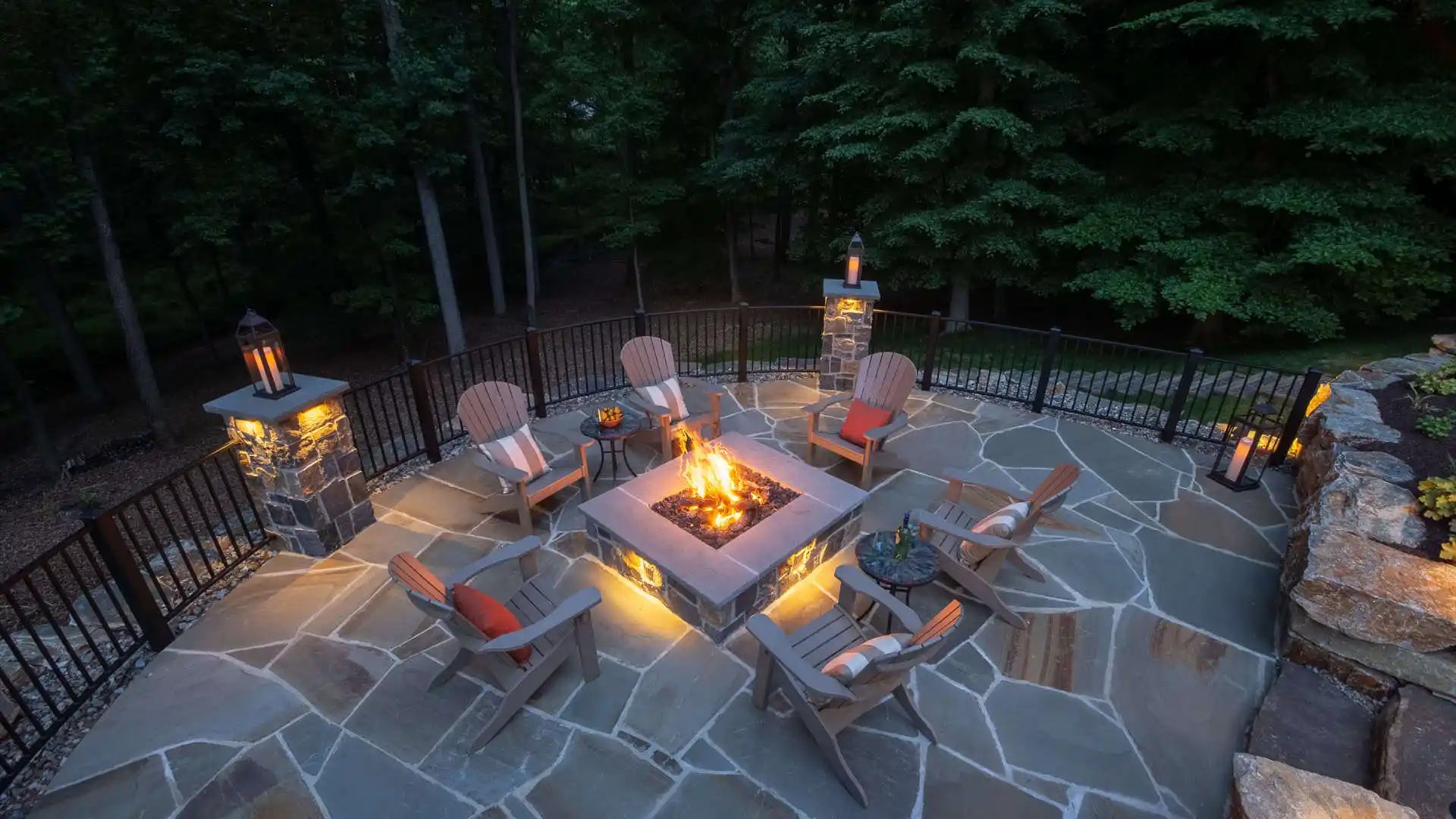 An outdoor patio in Delaware County, PA, surrounded by a metal railing, features a lit firepit and Adirondack chairs with cushions. Lanterns and stone flooring enhance the cozy ambiance, perfectly blending nature with expertly crafted hardscaping services for inviting outdoor living.