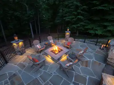 An outdoor patio in Delaware County, PA, surrounded by a metal railing, features a lit firepit and Adirondack chairs with cushions. Lanterns and stone flooring enhance the cozy ambiance, perfectly blending nature with expertly crafted hardscaping services for inviting outdoor living.