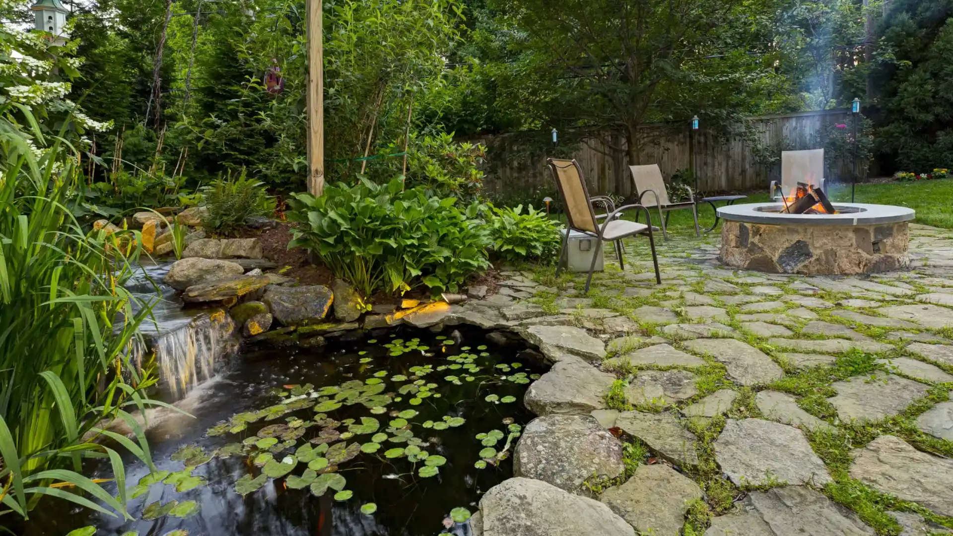 A serene backyard scene in Delaware County, PA, with a stone patio showcases the beauty of outdoor living. It features a smokey fire pit, two chairs, a lush garden, and a pond with lily pads and a small waterfall. Surrounded by trees, this tranquil spot offers an inviting retreat.