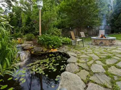 A serene backyard scene in Delaware County, PA, with a stone patio showcases the beauty of outdoor living. It features a smokey fire pit, two chairs, a lush garden, and a pond with lily pads and a small waterfall. Surrounded by trees, this tranquil spot offers an inviting retreat.