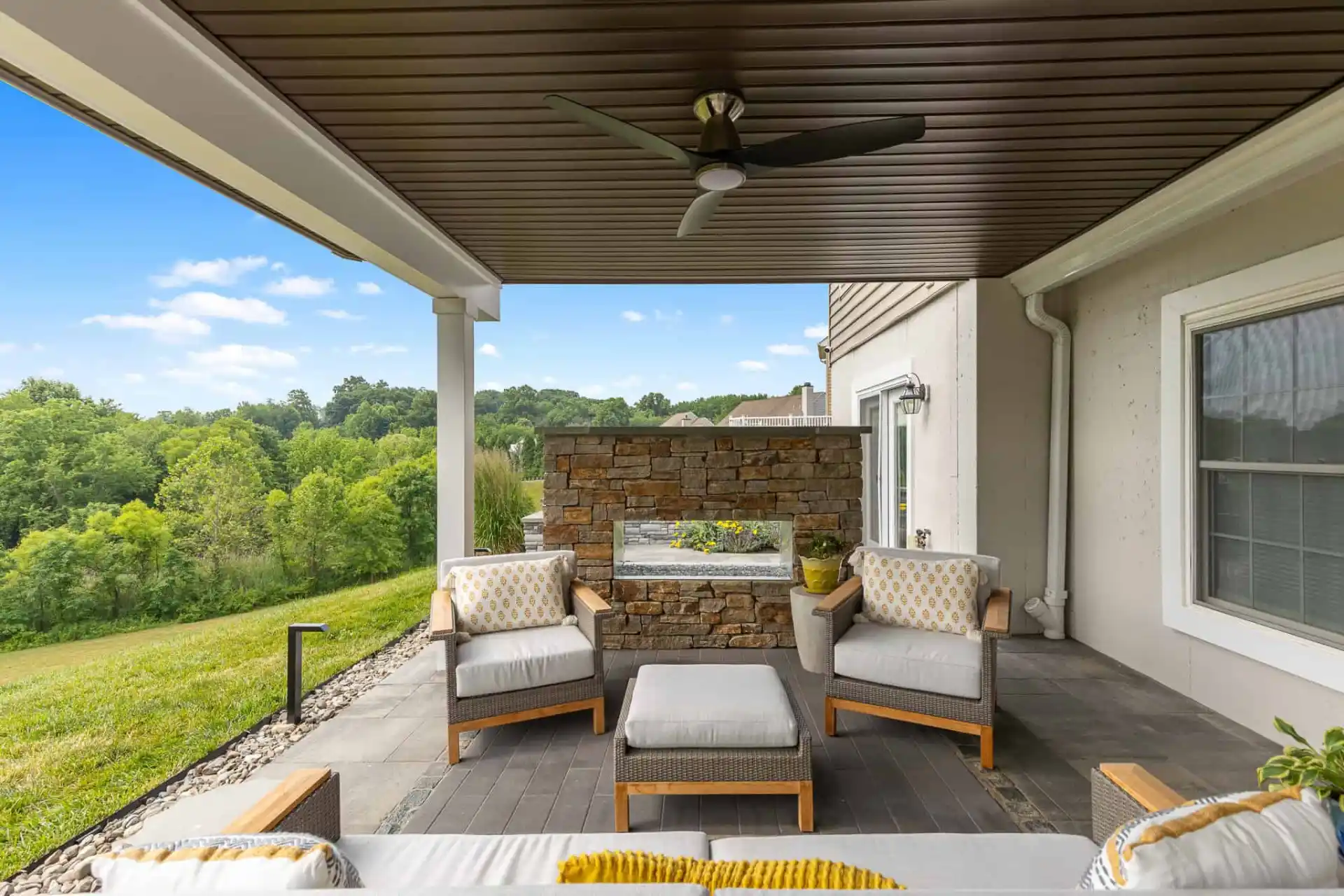 Covered patio with wooden furniture, including chairs and an ottoman with gray cushions. A ceiling fan is above, and a stone fireplace adds charm. This inviting outdoor living space in Delaware County, PA, overlooks a lush landscape with trees under a bright blue sky and scattered clouds.