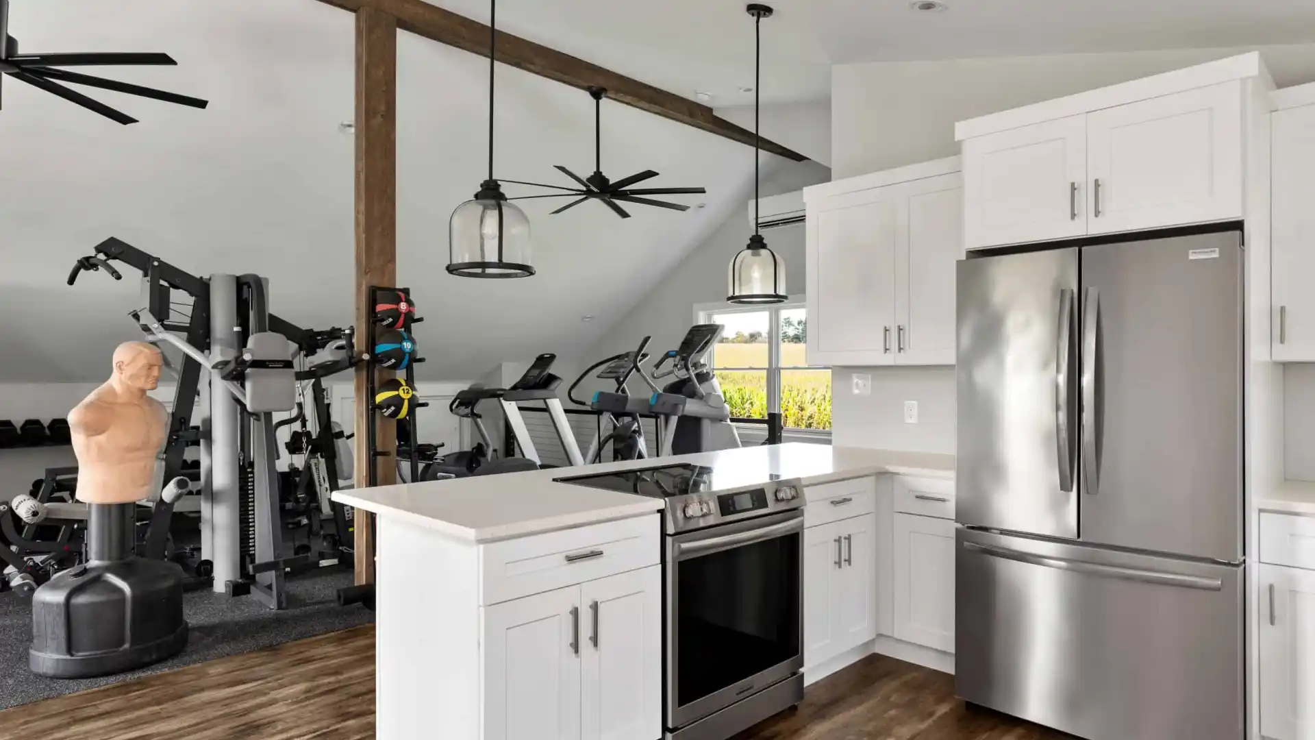 A modern open-concept space combines a kitchen and a gym, perfect for those who love indoor and outdoor living. The kitchen boasts white cabinets and stainless steel appliances, while the gym features exercise machines, weights, and a training dummy. A wooden beam and ceiling fans complete the look.