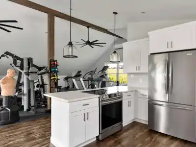 A modern open-concept space combines a kitchen and a gym, perfect for those who love indoor and outdoor living. The kitchen boasts white cabinets and stainless steel appliances, while the gym features exercise machines, weights, and a training dummy. A wooden beam and ceiling fans complete the look.