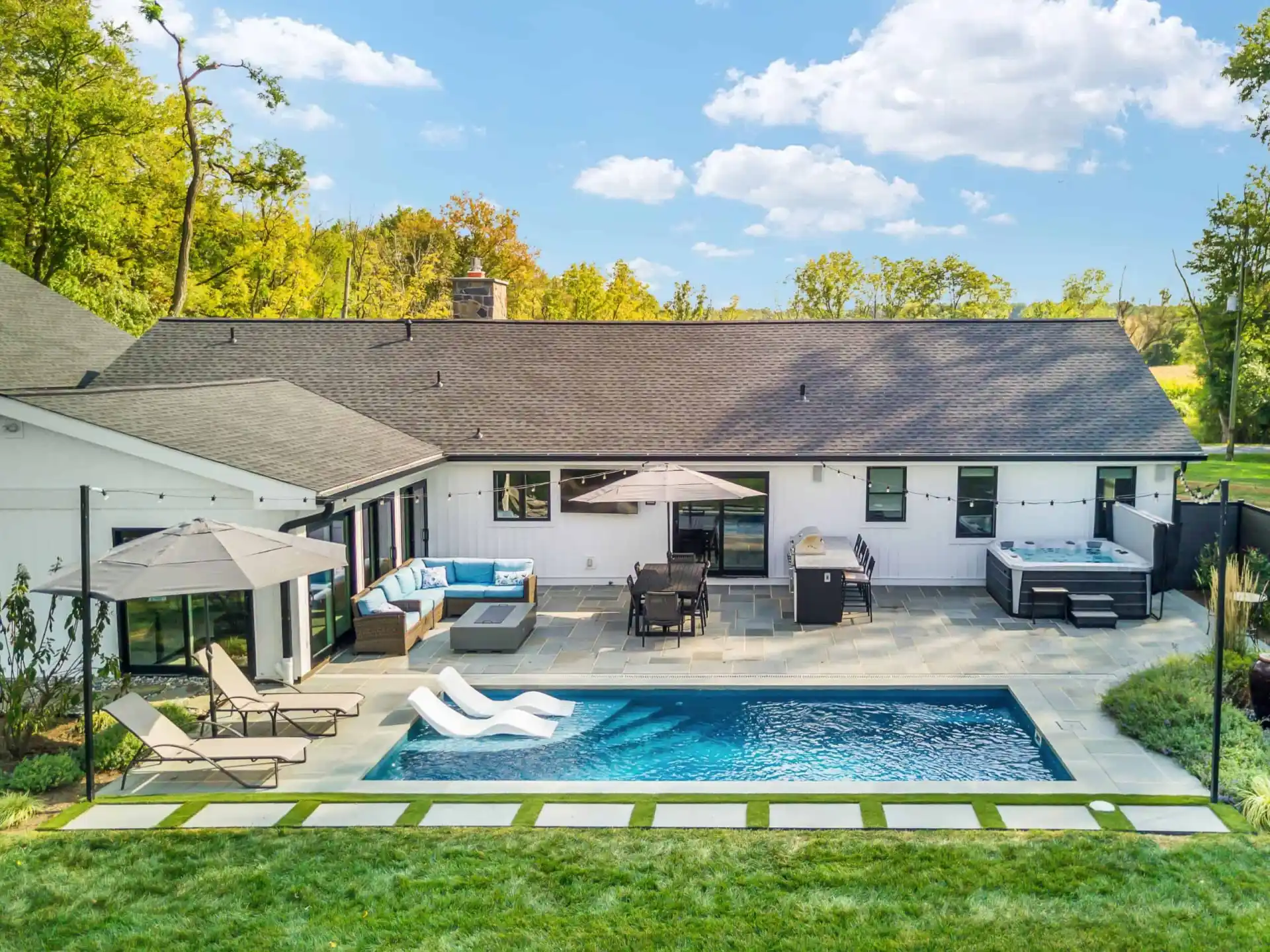 Aerial view of a modern backyard in Delaware County, PA, featuring stunning hardscaping services. A swimming pool is surrounded by a stone patio with lounge chairs, a dining set under an umbrella, a hot tub, and sectional sofa. The white house has a gray roof with trees in the background.