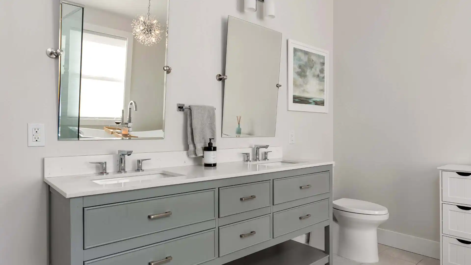 Modern bathroom with a double vanity featuring gray cabinets and two mirrors above. A white countertop with dual sinks and a sleek soap dispenser completes the look. To the right, find the toilet near a window adorned with blinds and wall art, echoing outdoor living vibes from Delaware County, PA.