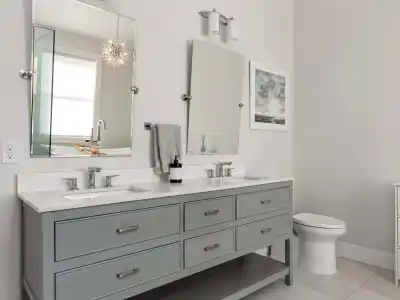 Modern bathroom with a double vanity featuring gray cabinets and two mirrors above. A white countertop with dual sinks and a sleek soap dispenser completes the look. To the right, find the toilet near a window adorned with blinds and wall art, echoing outdoor living vibes from Delaware County, PA.