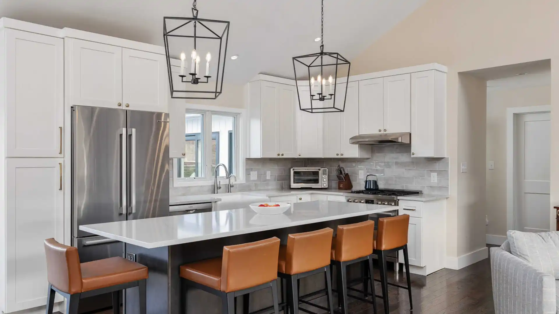 Modern kitchen with white cabinets, stainless steel appliances, and a gray island. Brown leather bar stools line the island beneath two geometric pendant lights. Wood flooring enhances this cozy space, seamlessly blending indoor elegance with outdoor living in Delaware County, PA.