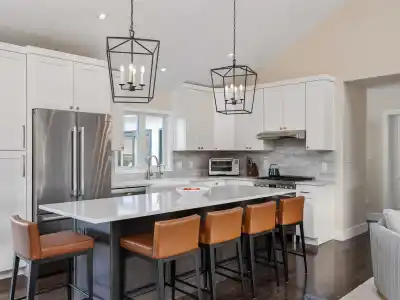 Modern kitchen with white cabinets, stainless steel appliances, and a gray island. Brown leather bar stools line the island beneath two geometric pendant lights. Wood flooring enhances this cozy space, seamlessly blending indoor elegance with outdoor living in Delaware County, PA.