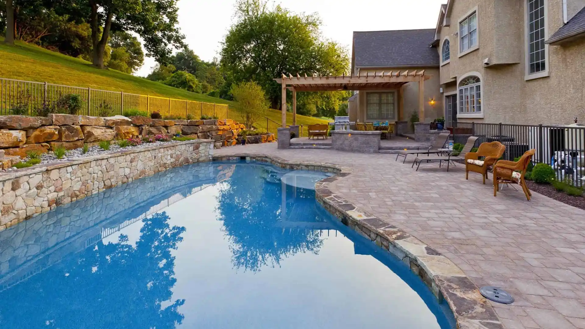 A luxurious backyard features a curved pool with a stone border, enhanced by expert landscaping services in Delaware County, PA. Surrounded by stylish patio furniture and a pergola with outdoor seating, the serene setting includes a stone wall lining the manicured garden.
