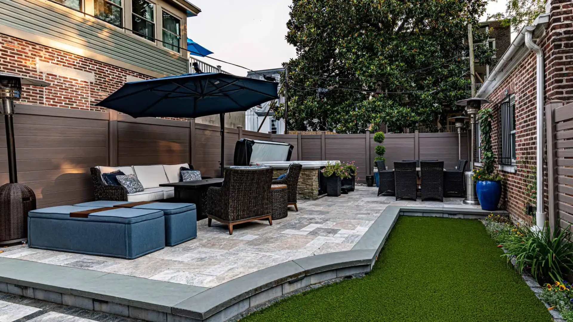 A spacious patio highlights outdoor living in Delaware County, PA, featuring wicker chairs and a large umbrella. A dining table sits nearby. The area is embraced by wooden fencing and a lush artificial grass lawn, framed by brick walls.