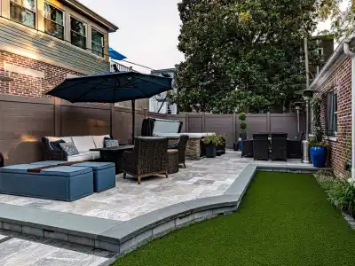 A spacious patio highlights outdoor living in Delaware County, PA, featuring wicker chairs and a large umbrella. A dining table sits nearby. The area is embraced by wooden fencing and a lush artificial grass lawn, framed by brick walls.