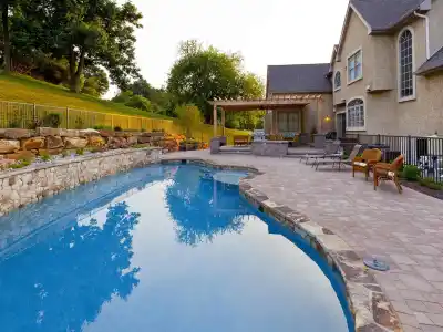 A luxurious backyard features a curved pool with a stone border, enhanced by expert landscaping services in Delaware County, PA. Surrounded by stylish patio furniture and a pergola with outdoor seating, the serene setting includes a stone wall lining the manicured garden.