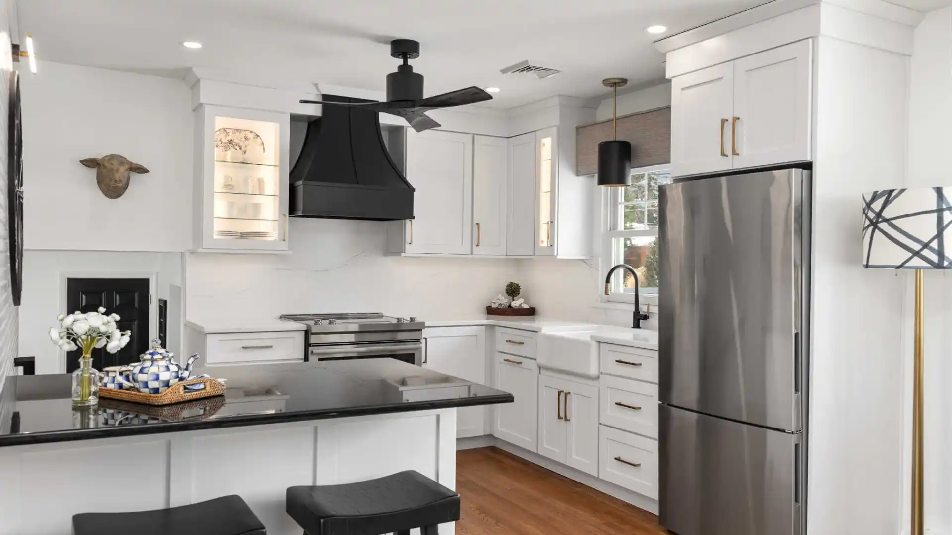 Modern kitchen with white cabinets, stainless steel appliances, a black range hood, and a farm-style sink. A basket of blue-patterned napkins sits on the black countertop island with two stools. Decorative wall art and a large pendant light add style, reflecting an ambiance reminiscent of elegant outdoor living in Delaware County, PA.