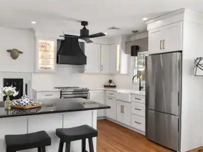 Modern kitchen with white cabinets, stainless steel appliances, a black range hood, and a farm-style sink. A basket of blue-patterned napkins sits on the black countertop island with two stools. Decorative wall art and a large pendant light add style, reflecting an ambiance reminiscent of elegant outdoor living in Delaware County, PA.