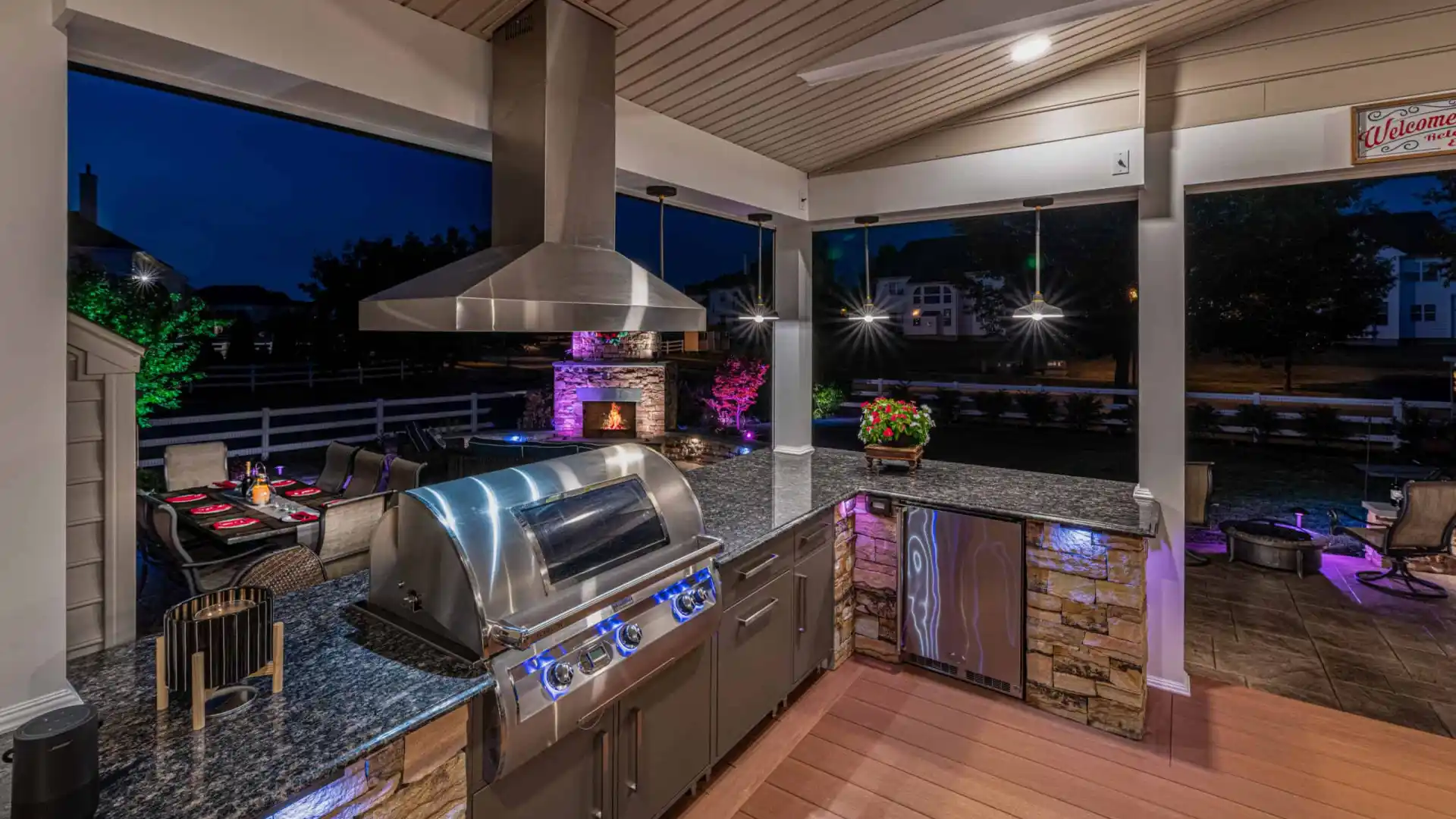 Outdoor living in Delaware County, PA is elevated with this nighttime kitchen, featuring stone countertops and a stainless steel grill. Ambient lighting highlights the area, while a fireplace and colorful patio lights create a cozy atmosphere. Perfectly executed by premier hardscaping services.