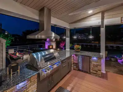 Outdoor living in Delaware County, PA is elevated with this nighttime kitchen, featuring stone countertops and a stainless steel grill. Ambient lighting highlights the area, while a fireplace and colorful patio lights create a cozy atmosphere. Perfectly executed by premier hardscaping services.