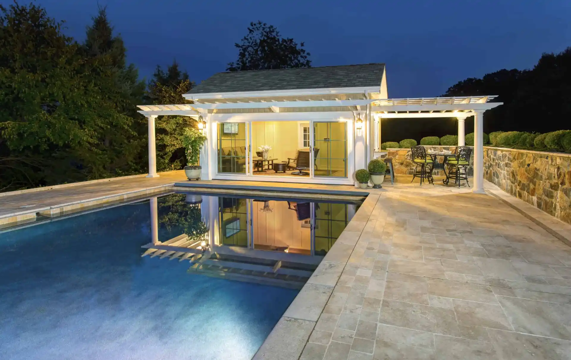 Elegant modern pool house illuminated at night with a pergola and glass doors. Surrounded by a beautifully designed patio and adjacent to a swimming pool, it reflects the structure. Outdoor furniture enhances the outdoor living vibe of Delaware County, PA, with trees visible in the background.