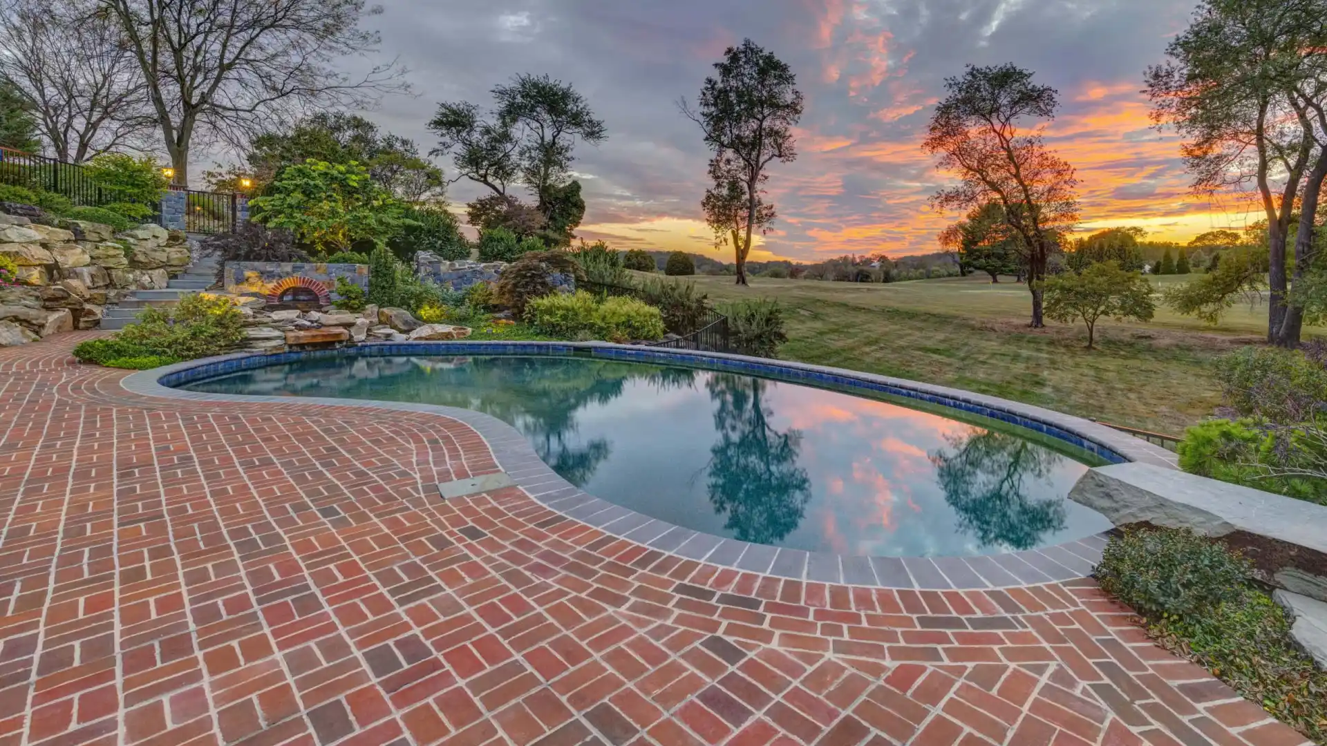 A serene swimming pool surrounded by a brick patio is pictured at sunset, showcasing outdoor living in Delaware County, PA. The sky, painted with hues of orange, pink, and purple, reflects on the water's surface. Trees and greenery border the area as a grassy lawn extends into the distance.