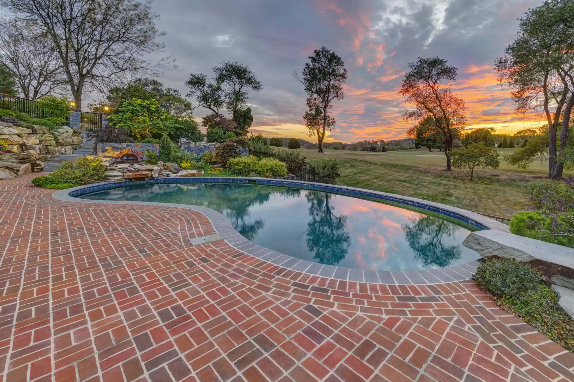 A serene swimming pool surrounded by a brick patio is pictured at sunset, showcasing outdoor living in Delaware County, PA. The sky, painted with hues of orange, pink, and purple, reflects on the water's surface. Trees and greenery border the area as a grassy lawn extends into the distance.