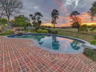 A serene swimming pool surrounded by a brick patio is pictured at sunset, showcasing outdoor living in Delaware County, PA. The sky, painted with hues of orange, pink, and purple, reflects on the water's surface. Trees and greenery border the area as a grassy lawn extends into the distance.