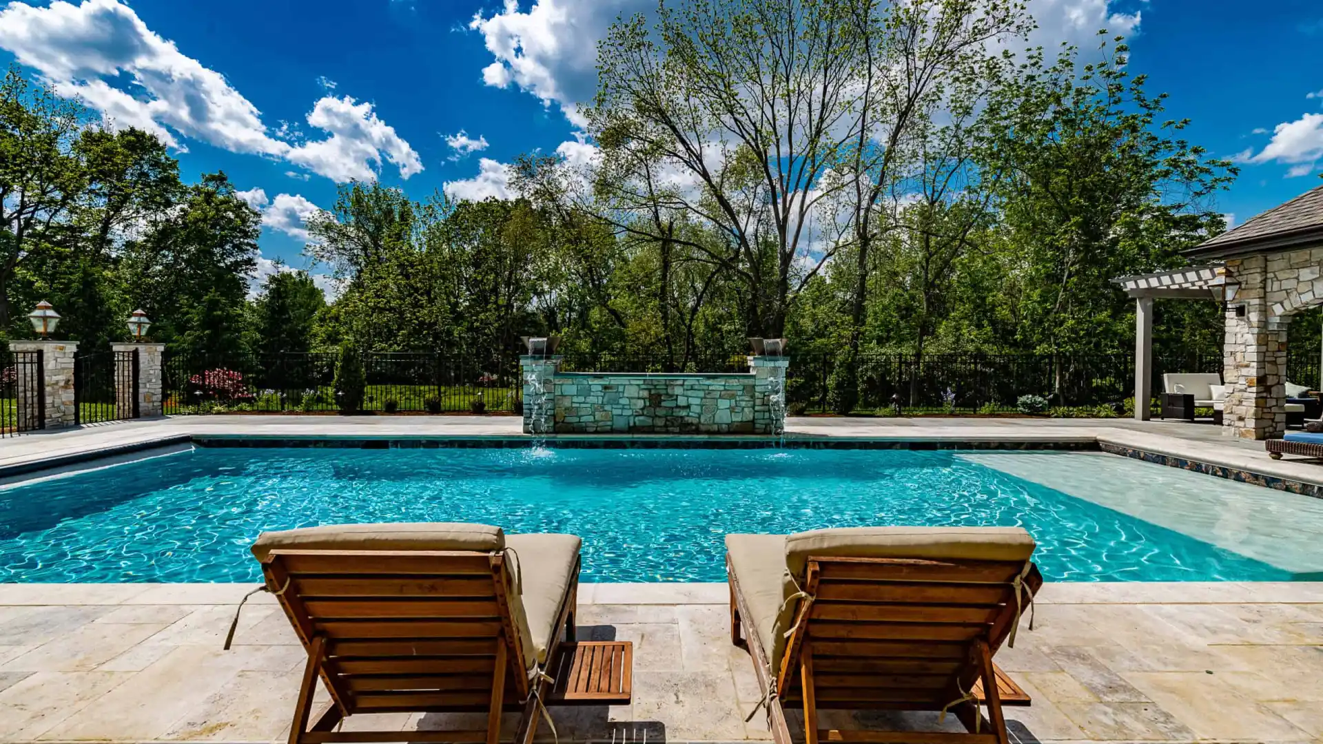 Two wooden lounge chairs face a pristine, rectangular swimming pool under a partly cloudy blue sky. The pool is surrounded by a stone patio and lush greenery, showcasing the finest hardscaping services Delaware County PA has to offer for an idyllic outdoor living experience.