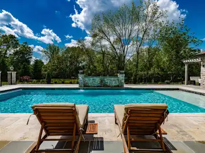 Two wooden lounge chairs face a pristine, rectangular swimming pool under a partly cloudy blue sky. The pool is surrounded by a stone patio and lush greenery, showcasing the finest hardscaping services Delaware County PA has to offer for an idyllic outdoor living experience.