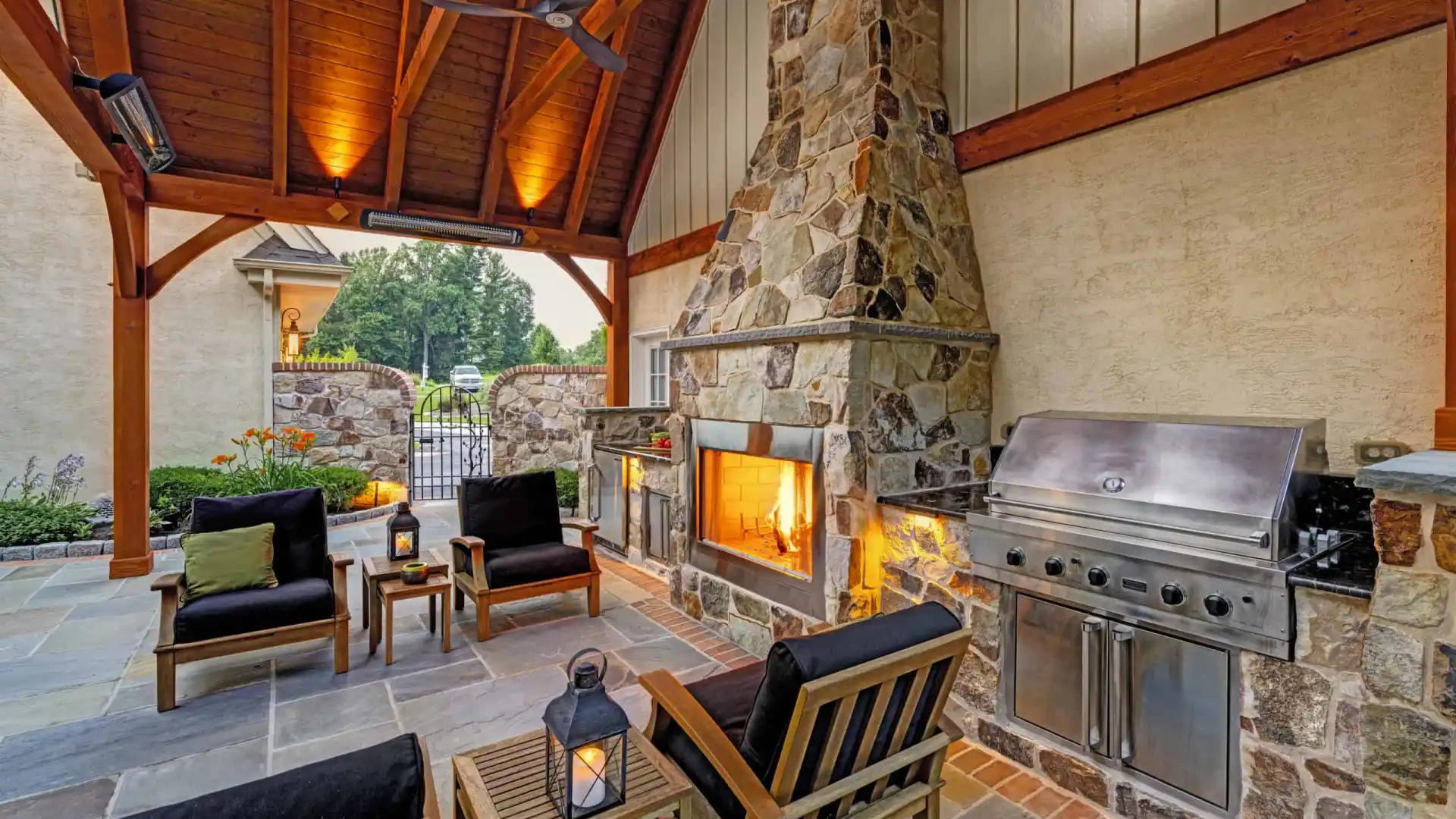 Outdoor patio featuring a stone fireplace with a fire, stainless steel grill, and wooden furniture with black cushions. Ceiling fan above and a lantern on the table. Stone tiled floor and garden visible in the background, perfect for enjoying outdoor living in Delaware County PA.