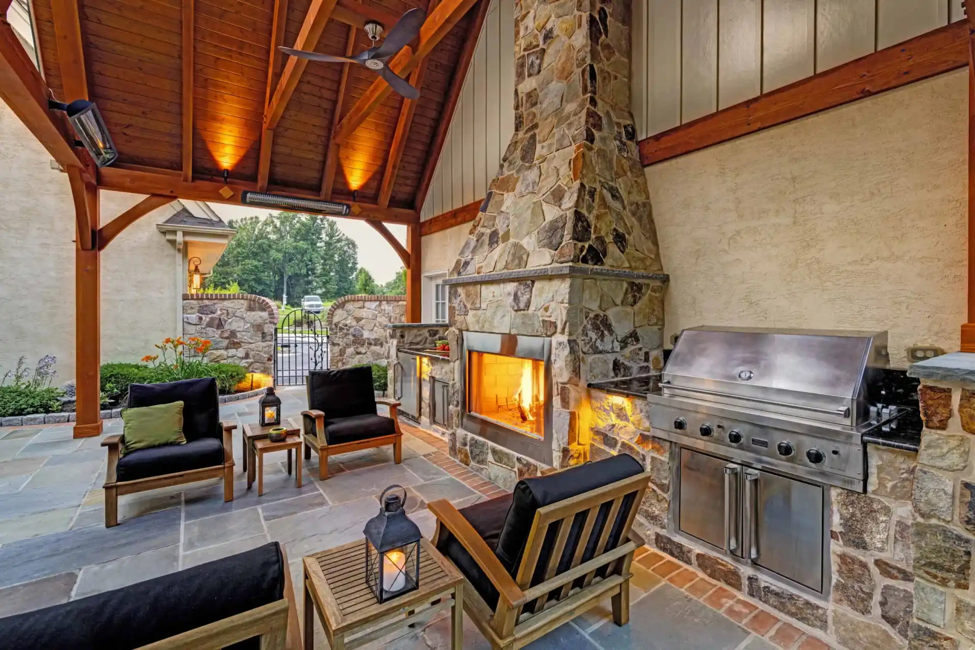 Outdoor patio featuring a stone fireplace with a fire, stainless steel grill, and wooden furniture with black cushions. Ceiling fan above and a lantern on the table. Stone tiled floor and garden visible in the background, perfect for enjoying outdoor living in Delaware County PA.
