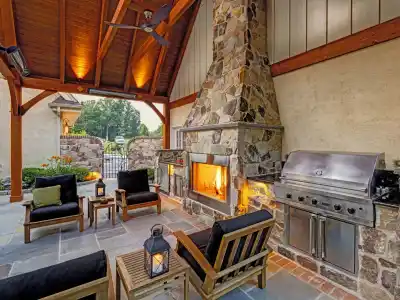 Outdoor patio featuring a stone fireplace with a fire, stainless steel grill, and wooden furniture with black cushions. Ceiling fan above and a lantern on the table. Stone tiled floor and garden visible in the background, perfect for enjoying outdoor living in Delaware County PA.