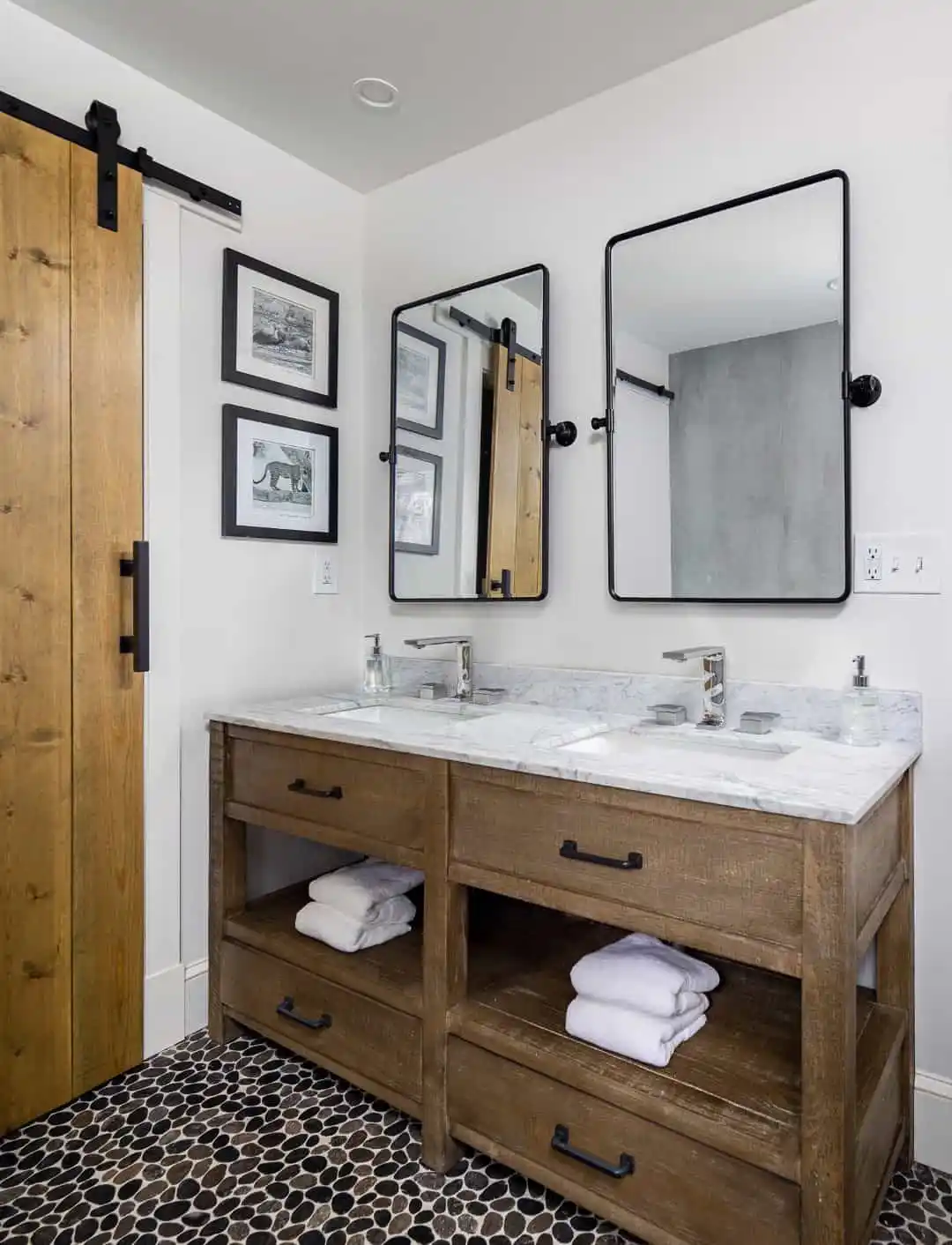 Modern bathroom with a wooden double vanity featuring a marble countertop and two black-framed mirrors. Towels rest on open shelves below. A sliding barn door and framed black and white photos adorn the walls, while the pebble-like floor pattern infuses an outdoor living aesthetic popular in Delaware County, PA.