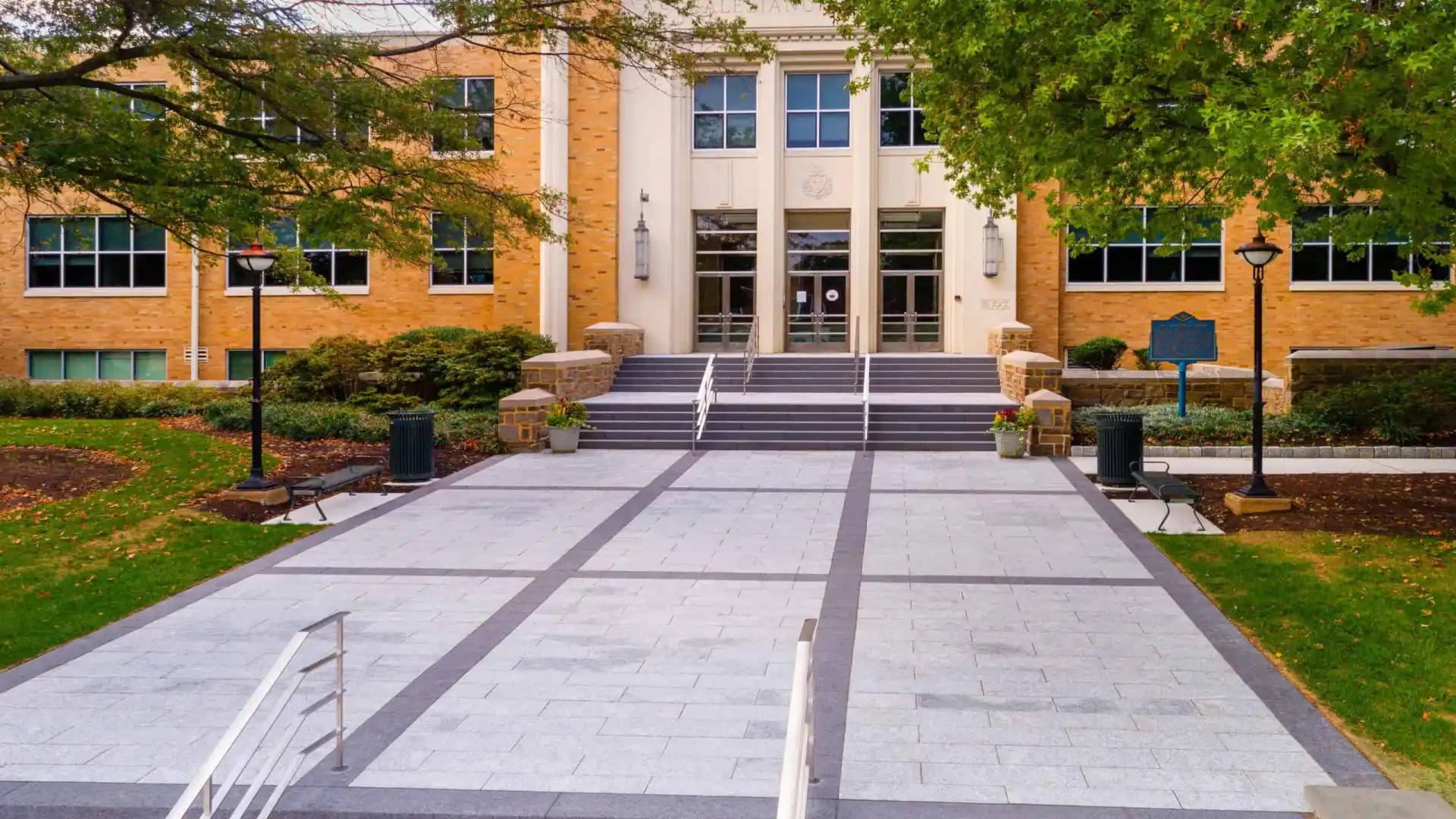 A brick academic building with large windows and white columns is surrounded by green trees, enhanced by expert landscaping services in Delaware County, PA. A wide stone walkway with steps leads to the entrance, featuring lamps on either side. The scene is bathed in soft daylight.
