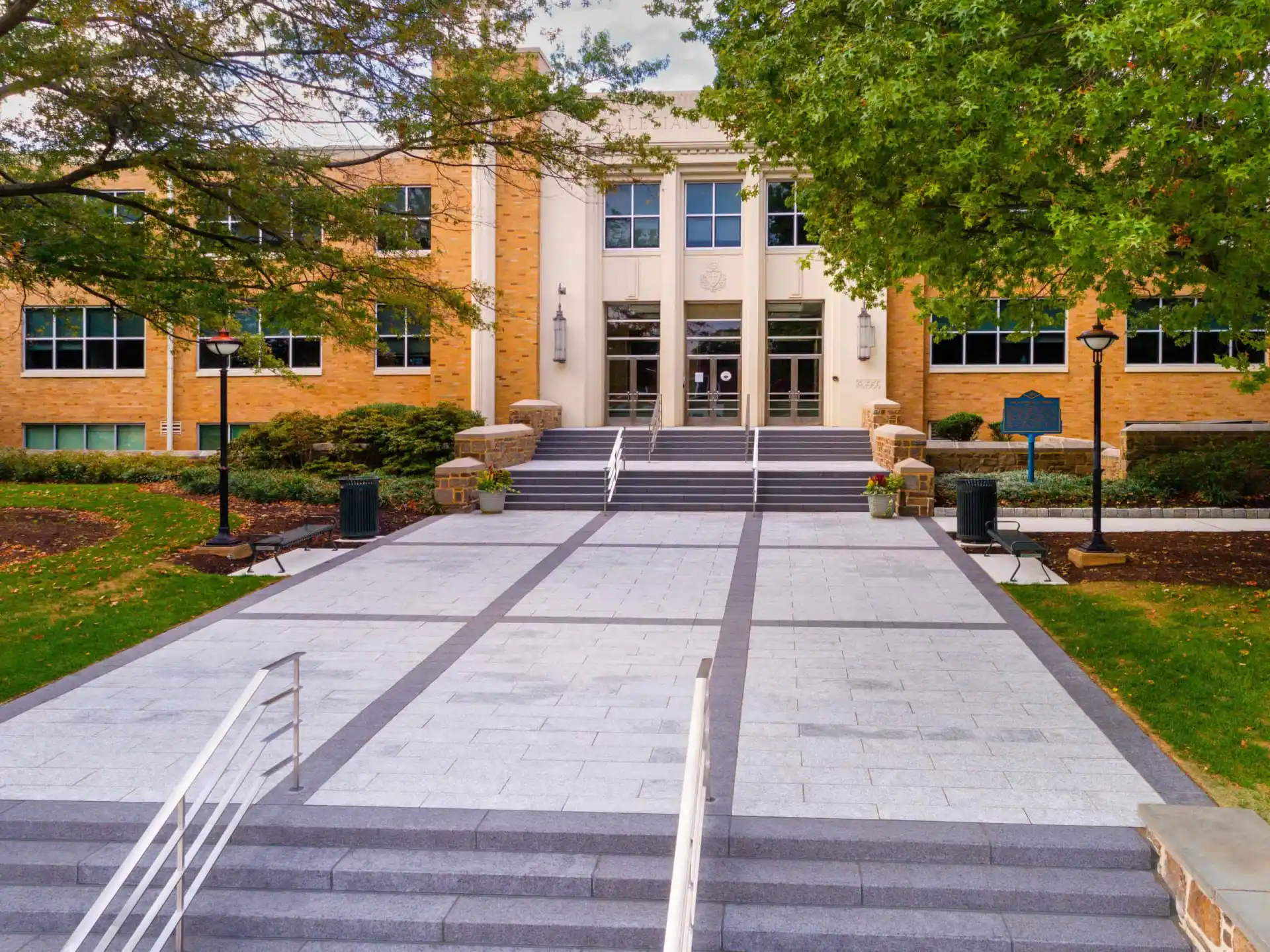 A brick academic building with large windows and white columns is surrounded by green trees, enhanced by expert landscaping services in Delaware County, PA. A wide stone walkway with steps leads to the entrance, featuring lamps on either side. The scene is bathed in soft daylight.