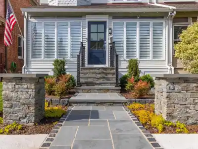 A front entrance with a stone walkway leads to steps and a blue door. Flanked by stone pillars and small shrubs, this Delaware County PA home showcases outdoor living with a sunroom featuring large windows. An American flag adorns the left, while neatly trimmed grass surrounds the path.