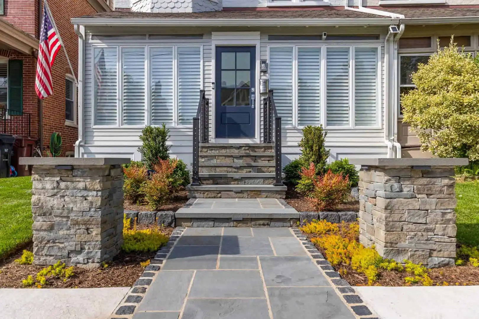 A front entrance with a stone walkway leads to steps and a blue door. Flanked by stone pillars and small shrubs, this Delaware County PA home showcases outdoor living with a sunroom featuring large windows. An American flag adorns the left, while neatly trimmed grass surrounds the path.