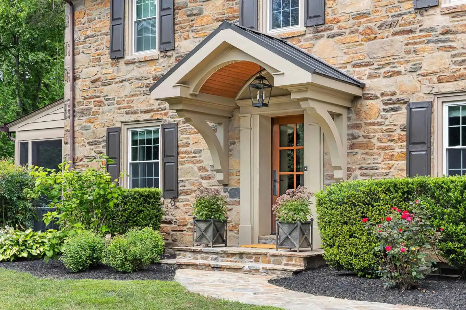 A stone house with a wooden front door under an arched portico invites you into refined outdoor living in Delaware County, PA. Black shutters and potted plants add charm, while neatly trimmed hedges and colorful flowers showcase expert landscaping services. A lantern hangs above the welcoming entry.
